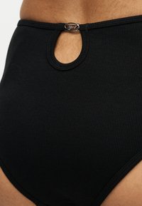 Sous-vêtement noir côtelé taille haute avec une petite découpe en forme de goutte et une agrafe argentée à l’avant de la ceinture, sur fond de peau.