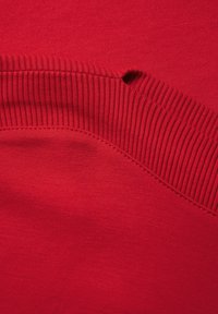 Lähikuva punaisesta kankaasta, jossa on uritettu reunus ja pieni reikä sauman lähellä, näyttäen tekstuuria ja ompeluyksityiskohtia.