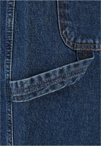 Denim tkanina z globoko modrim odtenkom ima majhen žep prišit na strani, poudarjen z oranžnim šivanjem in nekoliko zbledelim robom.