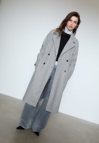 Grauer Wollmantel mit doppelreihigem Design, schwarzen Knöpfen, kombiniert mit einem schwarzen Oberteil, ausgestellten grauen Jeans und schwarzen spitzen Absätzen.