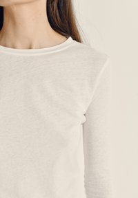 Žena u običnoj bijeloj majici dugih rukava, prikazana od vrata do sredine prsa na neutralnoj pozadini.