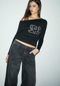 Schwarzes Off-Shoulder Langarmoberteil mit "Too Cute" Strassschriftzug, kombiniert mit weit geschnittenen schwarzen Jeans aus Denim. Glattes Material, figurbetontes Design.