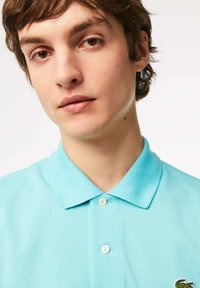 Polo shirt azzurro chiaro in tessuto testurizzato, con colletto e due bottoni, decorato con un piccolo logo verde sul lato sinistro.