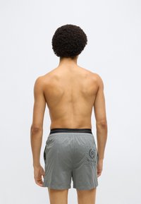 Szürke fürdőnadrág gumis fekete derékrésszel, hátul egy emblémás zsebbel és sima, könnyű anyagból.