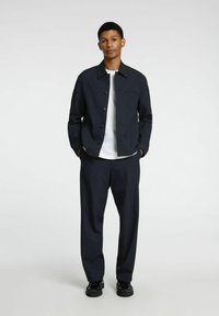Giacca navy con colletto piatto, chiusura a bottoni e taschino sul petto, abbinata a pantaloni a gamba larga coordinati e scarpe nere. Tessuto dalla trama liscia.