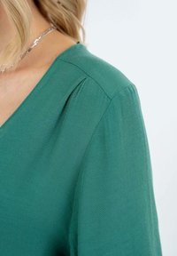 Žalia palaidinė su V iškirpte, trumpomis rankovėmis ir minkšta tekstūra su subtiliu blizgesiu. Klostuota detalė ant peties suteikia dimensijos.