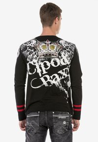 Schwarzes Langarmshirt mit weißen Flügeln, einer Krongrafik und dem Text "Cipoda Baxx", sowie roten Streifenakzenten an den Ärmeln.