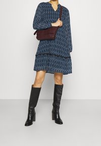 Marineblaues Kleid mit weißem Blattmuster, langen Ärmeln und gestuften Saum, kombiniert mit einer burgunderfarbenen Umhängetasche und knielangen schwarzen Stiefeln.
