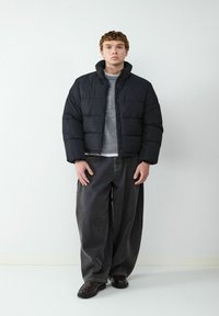 Czarna kurtka puchowa z wysokim kołnierzem, pikowane sekcje, noszona na szarym swetrze. Zestawiona ze luźnymi ciemnymi dżinsami i czarnymi butami.