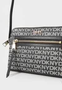 Borsa a tracolla DKNY bianca e nera con motivo a logo, dotata di una tasca con zip dorata, dettagli in pelle e una sottile tracolla regolabile.