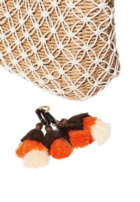 Sac en paille tissée avec motif textile blanc. Comprend trois pompons en orange, noir et crème attachés par un anneau en métal doré.