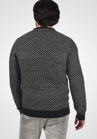 Uomo dai capelli scuri che indossa un maglione a righe bianche e nere e jeans grigi chiari, visto di spalle su uno sfondo bianco.