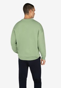 Felpa verde chiaro con una vestibilità rilassata, colletto a girocollo, polsini e orlo a coste, realizzata in morbido tessuto misto cotone. Indossata con pantaloni blu navy.