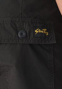 Un gros plan de tissu noir mettant en évidence un bouton et un patch logo "Stan Ray" jaune et noir. La texture semble lisse avec un motif subtil.