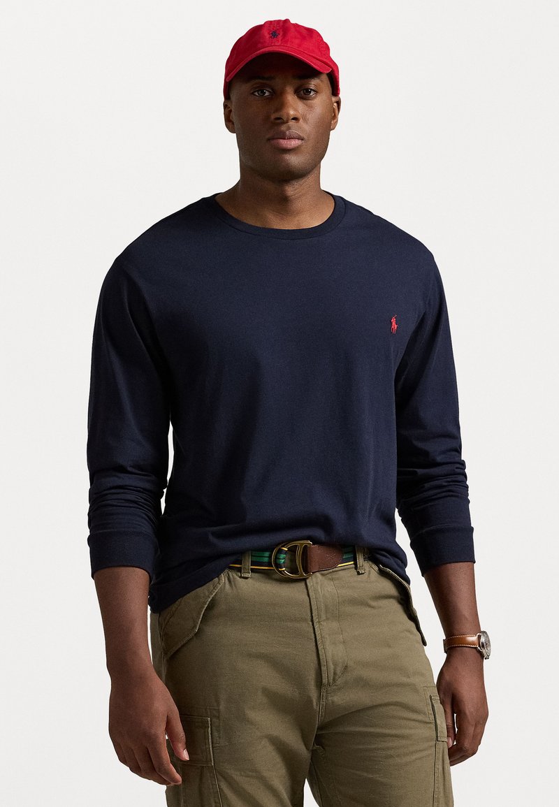 Navy langemouw T-shirt met ronde hals en een klein rood logo, gecombineerd met olijfgroene cargobroek en een rode pet, met een bruine riem en horloge.