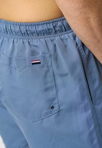 Šviesiai mėlyni šortai su elastingu juosmeniu, turi galinį kišenę ir mažą dryžuotą logotipo etikete raudonos, baltos ir mėlynos spalvų. Lygus paviršius.