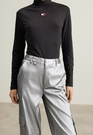 Personne portant un col roulé noir à manches longues avec le logo Tommy Jeans et un pantalon cargo argenté brillant, debout devant un fond neutre.