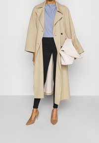 béžový trench coat s širokými rukávy, dvojitým zapínáním, nošený přes modré pruhované tričko a černé skinny kalhoty. Doplněný o texturovanou tašku a béžové podpatky.