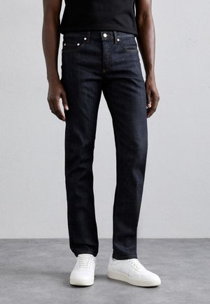Dark denim jeans with a straight leg fit, featuring copper stitching, five pockets, and a button fly, paired with white sneakers.