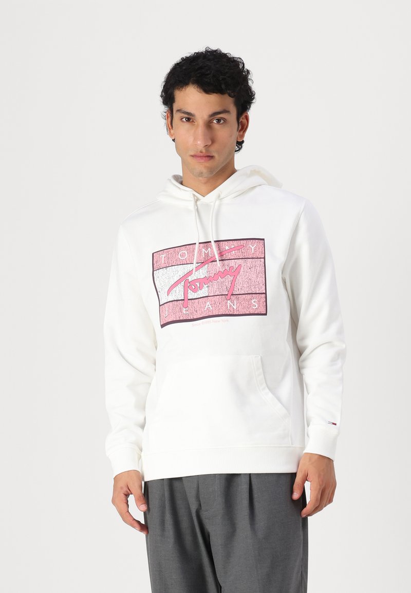 Hvit hettegenser med et stort rosa og grått grafisk logo av "Tommy Jeans" på forsiden. Har kengurulomme og snor i hetten.