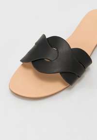 Zwarte leren sandaal met een gedraaide bandontwerp en een gladde beige binnenzool. De zool is lichtjes gestructureerd, wat de grip en stabiliteit verbetert.