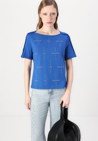 Blå kortermet T-skjorte med forhøyede tekstmønstre "LOVE" og "PATRIZIA PEPE." Kombinert med lyseblå jeans og en svart veske.