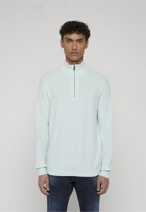 Mężczyzna z krótkimi kręconymi włosami, ubrany w jasnoniebieski sweter dzianinowy z długimi rękawami i kołnierzem na pół suwaka oraz ciemnoniebieskie dżinsy, stoi na jednolitym tle.