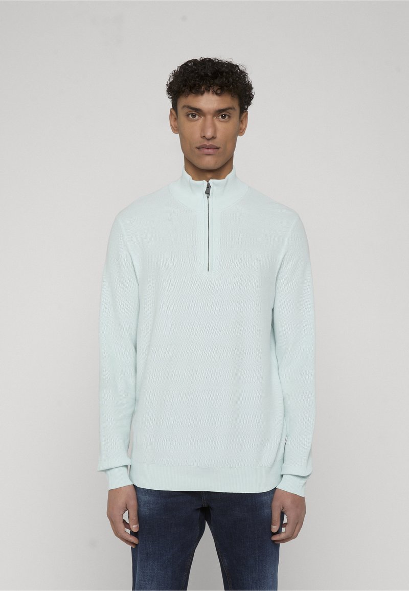 Homme aux cheveux courts et bouclés portant un pull léger bleu clair à manches longues avec col demi-fermeture éclair et jean bleu foncé, debout devant un fond uni.