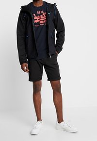 Chaqueta negra con cremallera y detalles azules, camiseta azul marino con gráfico rojo, pantalones cortos negros y zapatillas blancas. El modelo está de pie sobre un fondo liso.