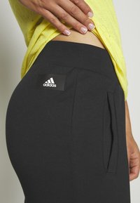 Schwarze Sporthosen mit Seitentaschen, aus einem glatten Stoff, mit einem Adidas-Logopatch am Bund. Kombiniert mit einem leuchtend gelben Shirt.