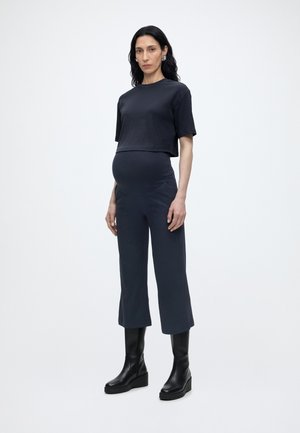 Tehotná žena stojaca, oblečená v čiernom krátkom rebrovanom topu, zladených nohaviciach a čiernych topánkach po polovicu lýtok, na jednoduchom svetlom pozadí.