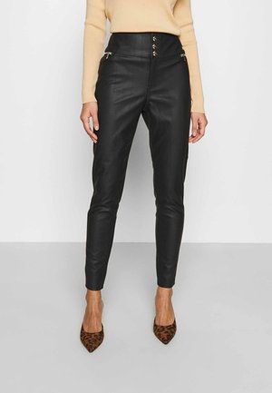 Femme portant un pantalon en cuir noir taille haute avec poches à fermeture éclair et trois boutons dorés, un pull beige et des talons pointus à imprimé léopard.