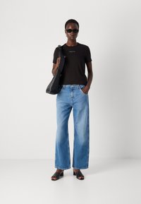 Svart figurnær T-skjorte med hvitt logo, lyseblå vide jeans, svart skulderveske, svarte blokkhælte sandaler og svarte solbriller.
