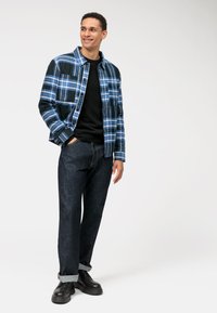 Blauwe geruite jas met donkere accenten, zwart sweatshirt en donkere spijkerbroek. Zwarte schoenen met een stevige zool. Eenvoudig ontwerp en casual pasvorm.