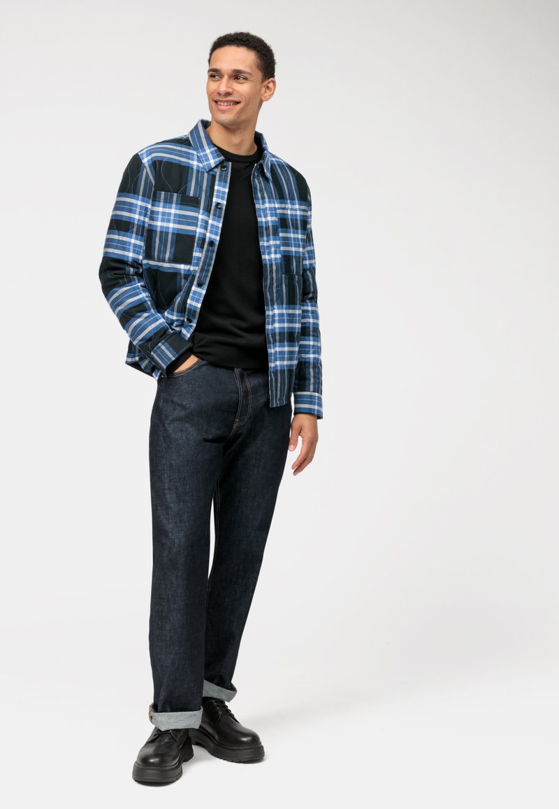 Blauwe geruite jas met donkere accenten, zwart sweatshirt en donkere spijkerbroek. Zwarte schoenen met een stevige zool. Eenvoudig ontwerp en casual pasvorm.