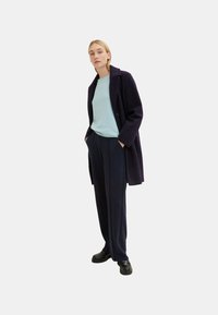 Navy blue wool coat with a textured finish, worn over a light blue sweater, paired with tailored black trousers and sturdy shoes.