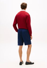 Κόκκινο φούτερ με λευκό γιακά, σε συνδυασμό με ναυτικό σορτς. Μαύρα loafers ολοκληρώνουν την εμφάνιση. Ομαλή υφή και casual σχεδιασμός.