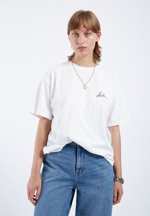 Weißes Baumwoll-T-Shirt mit lockerer Passform, kurzen Ärmeln, rundem Ausschnitt und einem kleinen schwarzen gestickten Detail auf der linken Brust.