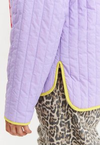 Fioletowa pikowana kurtka z żółtymi wykończeniami noszona na spodniach w panterkę, z widocznymi rękawami i zakrzywionym rozcięciem po bokach w okolicy biodra.