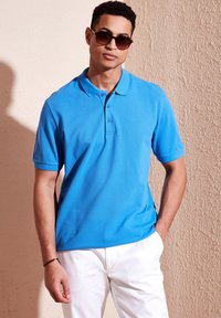 Polo de color azul claro con cuello, tapeta de tres botones y mangas cortas, combinado con pantalones blancos. Textura de tela suave.