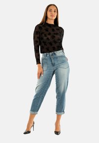 Top negro de manga larga con estampado floral; jeans de cintura alta desgastados en azul claro; puños doblados; tacones altos negros. Fondo minimalista.