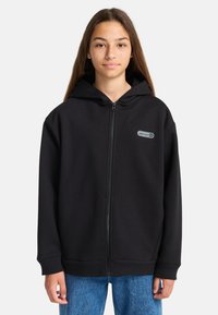 Adolescente che indossa una felpa nera con zip e un piccolo logo "element" sul petto, insieme a jeans blu, in piedi contro uno sfondo semplice.