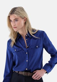 Satin blue button-up shirt with two chest pockets, golden buttons, and a collar. The model wears a brown belt with a rectangular buckle.