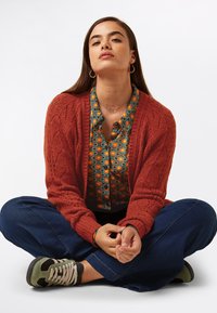 En stickad rostfärgad cardigan, mönstrad grön och orange skjorta under, avslappnade blå jeans och grå sneakers, sitter med benen i kors på en vit bakgrund.