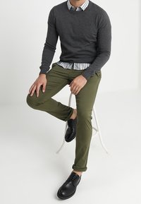 Dunkelgrauer Pullover mit Rundhalsausschnitt über einem weiß-blau gestreiften Hemd, kombiniert mit olivgrünen Chinos und schwarzen Schnürschuhen.