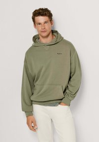 Olivno zelena jopa s kapuco in sprednjim žepom ter rebrastimi manšetami, na prsih z diskretno nalepko. Nošena čez sivo majico s kratkimi rokavi.