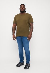 Oliven grønn kortermet henley-skjorte med tre knapper; kombinert med smale blå jeans og svarte sportssko. Glatt stoff, avslappet passform.