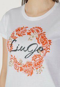 Weißes Baumwoll-T-Shirt mit einem aufwendigen orange-beigen Paisleymuster, in dessen Mitte der Name "Liu Jo" in einer dunklen, strukturierten Schriftart steht.