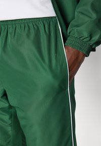 Green sports tracksuit featuring a textured fabric, elastic waistband, and contrasting white side piping. Close-up of pants with hand in pocket.
