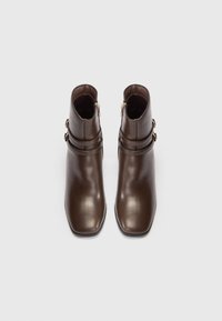 Par de botas de cuero marrón por el tobillo con puntas cuadradas y correas laterales decorativas con hebillas, vistas desde arriba sobre un fondo blanco.